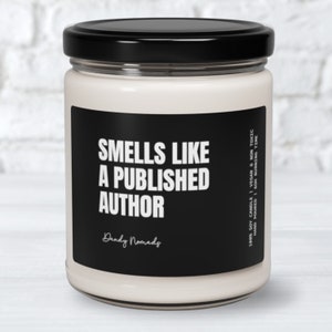 May include: A black and white candle with the text "Smells Like A Published Author" and the brand name "Dandy Nomads". The candle is in a clear glass jar with a black lid.