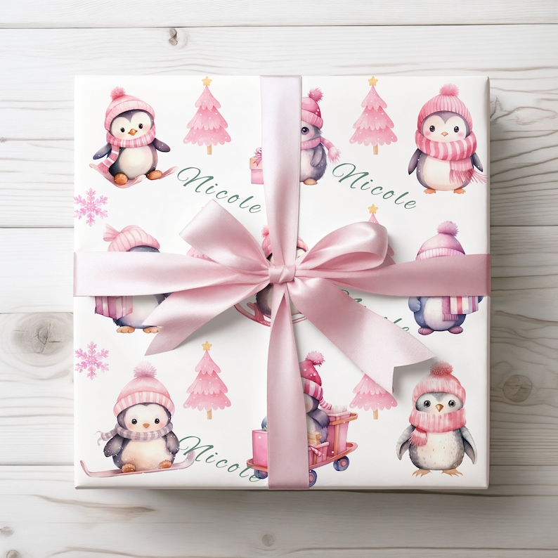 May include: A wrapped gift with a white background featuring a repeating pattern of penguins in pink hats and scarves, pink Christmas trees, and snowflakes. The gift is tied with a pink satin ribbon and bow. The name "Nicole" is written in green cursive.