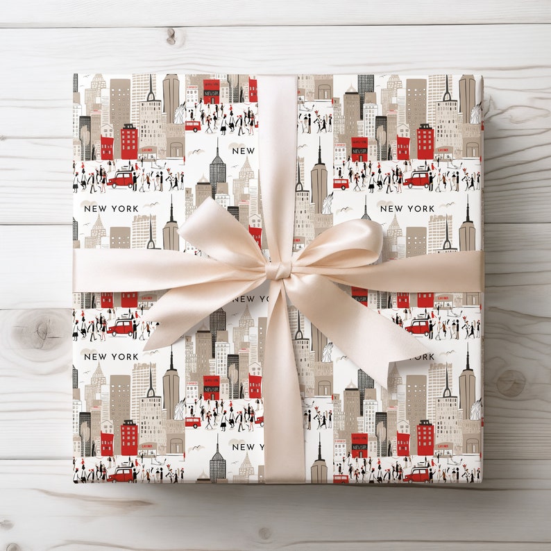 May include: A white gift wrap with a repeating pattern of a city skyline, red buses, and the text "NEW YORK". The gift wrap is tied with a cream-colored satin ribbon.