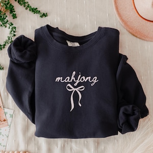 May include: A navy blue sweatshirt with the word "mahjong" in a cursive font and a bow graphic in white. The sweatshirt is folded, with the sleeves rolled up. The background includes a hat and greenery.