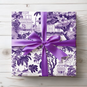 Puede incluir: Una caja de regalo envuelta en papel blanco con un patrón floral morado y blanco. La caja está atada con un gran lazo de satén morado.