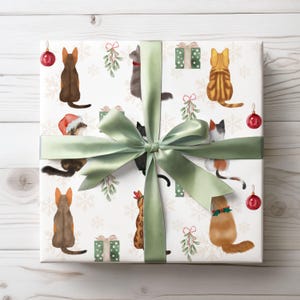 K&ouml;nnte beinhalten: Ein verpacktes Geschenk mit wei&szlig;em Hintergrund, das Aquarellillustrationen von Katzen, Schneeflocken, Mistelzweigen und kleinen gr&uuml;nen Geschenkboxen zeigt. Das Geschenk ist mit einem salbeigr&uuml;nen Band und einer Schleife gebunden, und rote Weihnachtskugeln sind sichtbar.