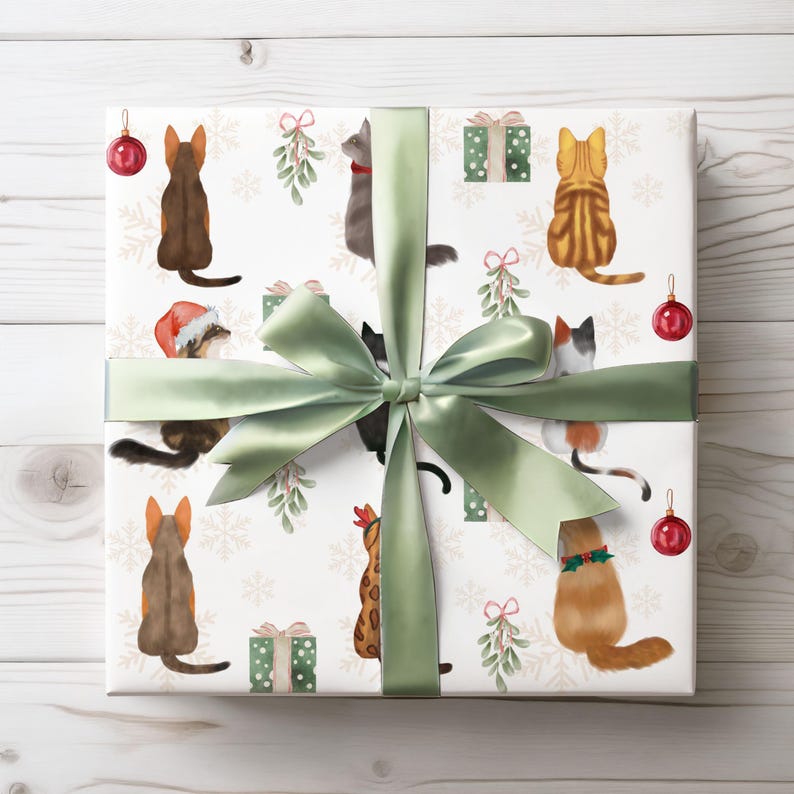 K&ouml;nnte beinhalten: Ein verpacktes Geschenk mit wei&szlig;em Hintergrund, das Aquarellillustrationen von Katzen, Schneeflocken, Mistelzweigen und kleinen gr&uuml;nen Geschenkboxen zeigt. Das Geschenk ist mit einem salbeigr&uuml;nen Band und einer Schleife gebunden, und rote Weihnachtskugeln sind sichtbar.