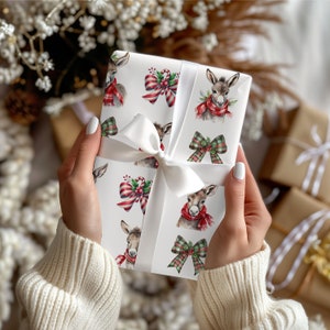 May include: A wrapped gift with white ribbon. The wrapping paper features a pattern of donkeys wearing red scarves, and red and green bows. The gift is held by a person wearing a cream-coloured sweater. Other gifts are in the background.