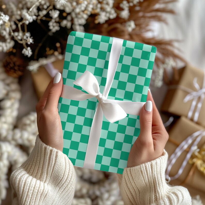 Op de afbeelding: Een groene en witte geruite cadeaudoos met een wit satijnen lint dat in een strik is gebonden. De doos wordt vastgehouden door een persoon die een witte trui draagt.