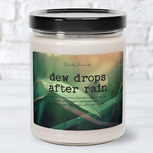 May include: A cream-colored candle in a glass jar with a black lid. The label reads "dew drops after rain" with a green leaf design. The candle is a soy wax blend with a 40-50 hour burn time.
