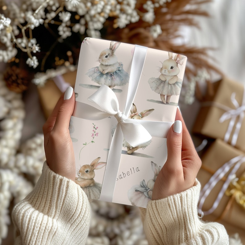 May include: A pink gift wrap with a white ribbon and a pattern of bunnies wearing tutus. The text "Isabella" is written on the wrapping paper.