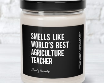 May include: A white candle in a glass jar with a black lid. The candle has a black label with the text "Smells Like World's Best Agriculture Teacher" and the brand name "Dandy Nomads". The candle is 100% soy, vegan, and non-toxic.