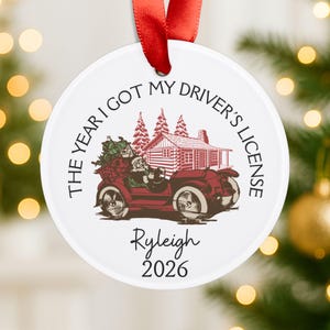 Pu&ograve; includere: Un ornamento rotondo in ceramica bianca con un nastro rosso. Il design presenta un'auto d'epoca rossa guidata da Babbo Natale, una baita in legno e alberi di pino. Il testo recita: THE YEAR I GOT MY DRIVER'S LICENSE, Ryleigh, 2026.