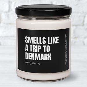 May include: A white candle with a black label that reads "Smells Like A Trip To Denmark" with the text "Dandy Nomads" below. The candle is in a glass jar with a black lid.