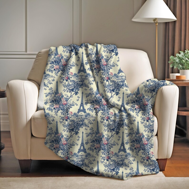 May include: A soft, cream-colored throw blanket with a repeating pattern of the Eiffel Tower and blue floral designs.