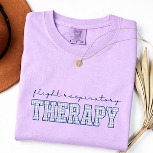 May include: A light purple t-shirt with the words "flight respiratory THERAPY" in blue script and block letters. The block letters have a blue and white gingham pattern. A gold necklace with a pendant is on the shirt.