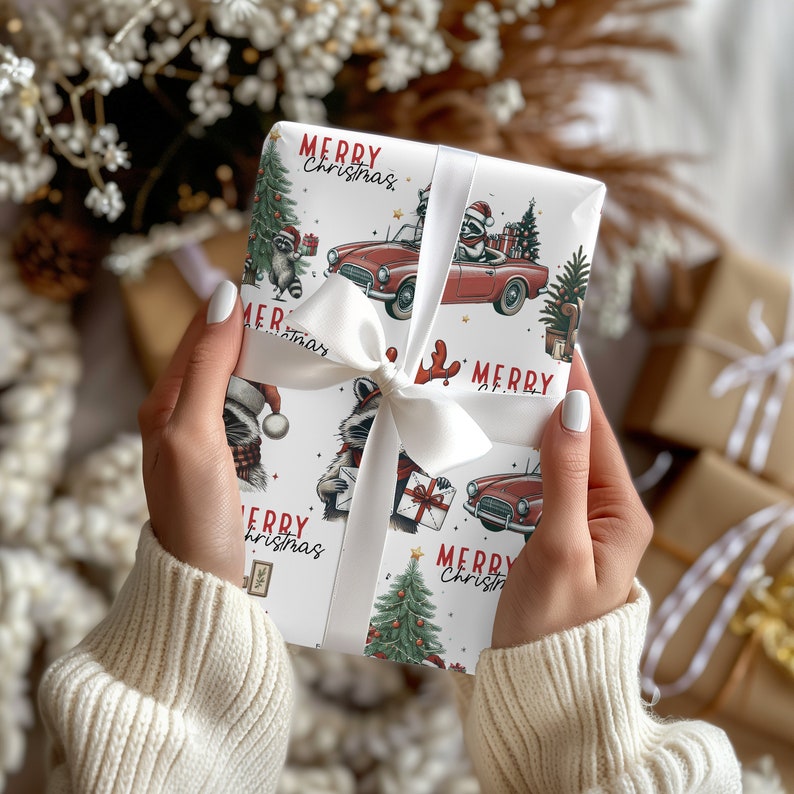 May include: A wrapped gift with white ribbon features a festive Christmas design. The wrapping paper has a white background with illustrations of raccoons, red cars, Christmas trees, and the words "Merry Christmas" in red. The gift is tied with a white satin ribbon.