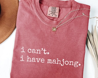 May include: A dusty rose Comfort Colors t-shirt with the text "i can't. i have mahjong." A silver necklace with a small pendant is draped across the shirt. The shirt is folded on a white surface.