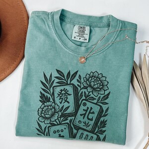 以下が含まれることがあります： 麻雀牌と花の黒いグラフィックが特徴の、シーフォームグリーンのTシャツ。Tシャツはクルーネックと半袖です。ペンダント付きのシルバーネックレスがTシャツの上にかけられています。Tシャツは白い面に折りたたまれています。