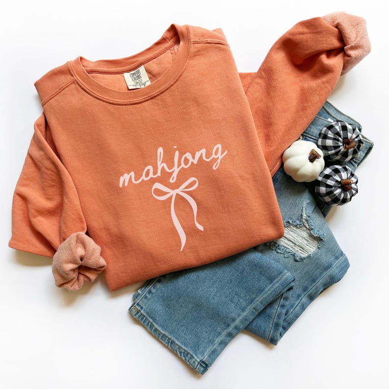 May include: An orange crewneck sweatshirt with the word "mahjong" in cursive script and a bow graphic. The sweatshirt is paired with light blue denim jeans and decorative pumpkins. The sleeves have a peach-colored cuff.