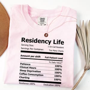 Peut inclure: T-shirt rose clair avec l'inscription "Residency Life" en noir. Le t-shirt présente un design de type tableau nutritionnel, indiquant les valeurs quotidiennes pour la patience, les heures cliniques, la privation de sommeil, la consommation de café, la cartographie, la résilience et le dévouement.