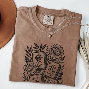 以下が含まれることがあります： 中国語の文字と花のモチーフが描かれた、茶色のComfort Colors Tシャツ。シルバーのネックレスがTシャツの上にかけられています。サイズS。