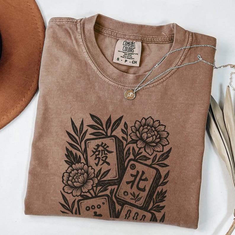 以下が含まれることがあります： 中国語の文字と花のモチーフが描かれた、茶色のComfort Colors Tシャツ。シルバーのネックレスがTシャツの上にかけられています。サイズS。