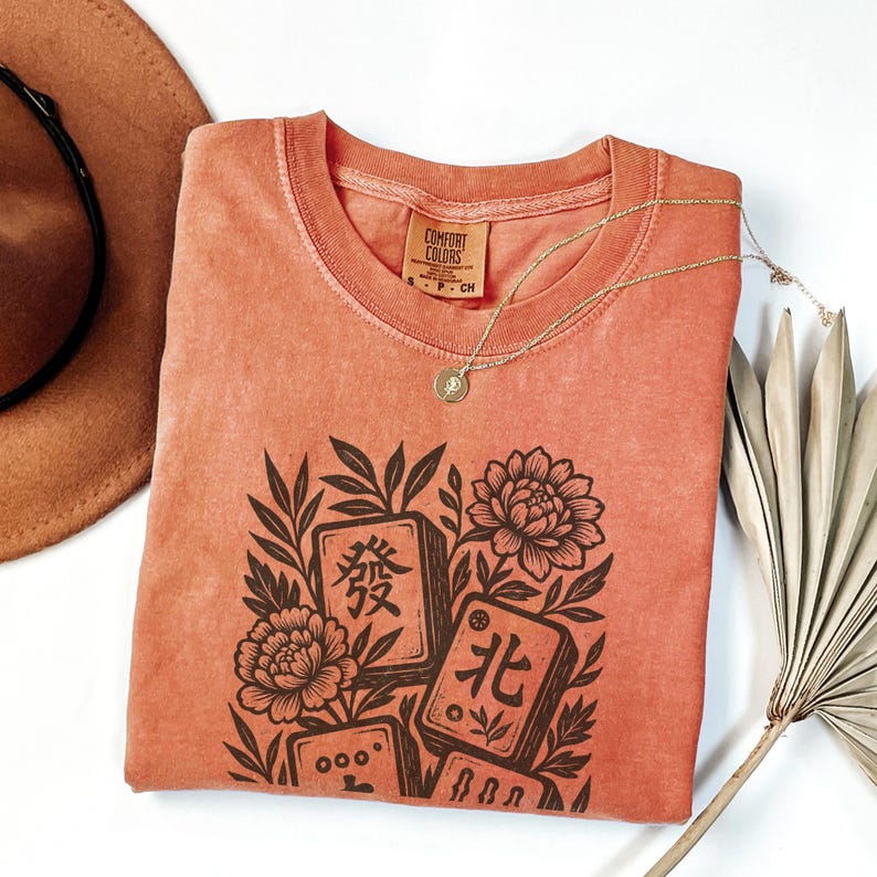 以下が含まれることがあります： コーラル色のComfort Colors Tシャツ。茶色の麻雀牌、花、葉のグラフィックが描かれています。 金色のネックレスがTシャツの上にあります。 茶色の帽子と乾燥したヤシの葉が背景にあります。