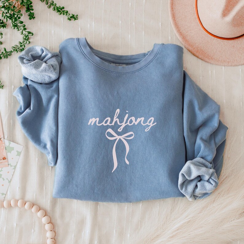 May include: A light blue sweatshirt with the word "mahjong" in a cursive font and a bow graphic. The sleeves are cuffed. A hat and greenery are in the background.