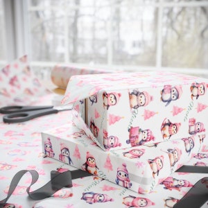 May include: Two gift boxes wrapped in white paper with a pattern of penguins wearing pink hats and scarves, and pink Christmas trees. The boxes are on a table with matching wrapping paper, scissors, and a black ribbon.