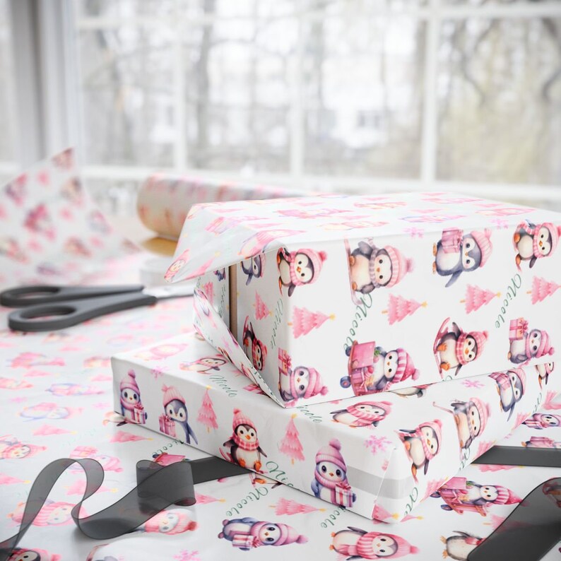 May include: Two gift boxes wrapped in white paper with a pattern of penguins wearing pink hats and scarves, and pink Christmas trees. The boxes are on a table with matching wrapping paper, scissors, and a black ribbon.