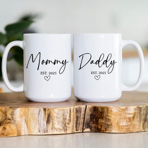 May include: Two white ceramic mugs with black lettering. The mug on the left says "Mommy Est. 2025" with a small heart. The mug on the right says "Daddy Est. 2025" with a small heart.