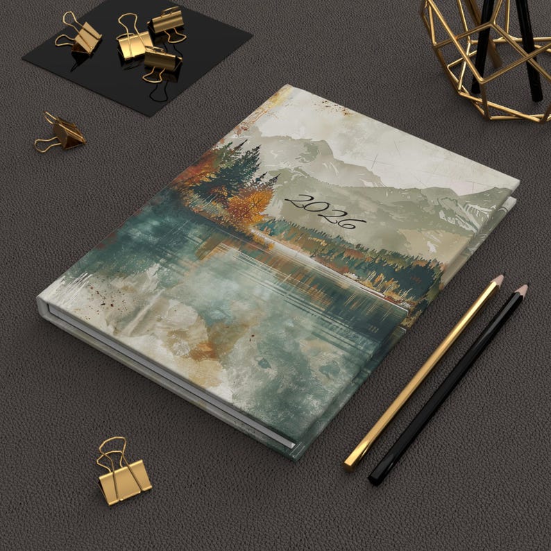 K&ouml;nnte beinhalten: Ein Hardcover-Journal mit einem Landschaftsgem&auml;lde eines Sees und Bergen, mit dem Jahr "2026" auf dem Einband. Goldene und schwarze Bleistifte und goldene B&uuml;roklammern sind ebenfalls vorhanden.