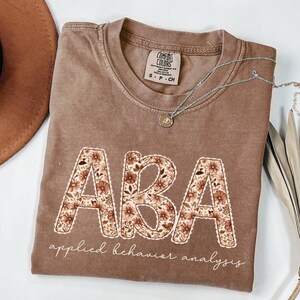 May include: A brown Comfort Colors t-shirt featuring the letters "ABA" in a floral pattern. The words "applied behaviour analysis" are written below the letters in a script font. A silver necklace with a pendant is draped across the shirt.