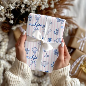 May include: A wrapped gift with pink and white striped paper featuring blue illustrations of lanterns, fans, and the word "mahjong." The gift is tied with a white satin ribbon and bow. Other wrapped gifts are in the background.