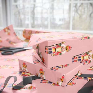 May include: Two wrapped gift boxes on a table, covered in pink wrapping paper with a repeating pattern of colorful nutcracker figures. A roll of matching paper, scissors, and black ribbon are also visible.