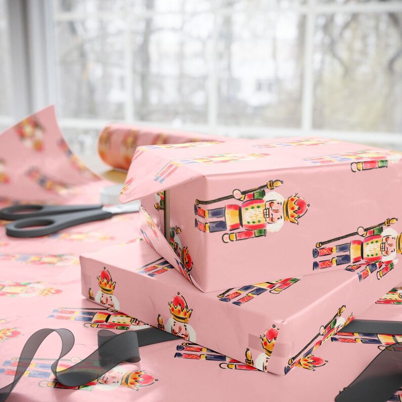 May include: Two wrapped gift boxes on a table, covered in pink wrapping paper with a repeating pattern of colorful nutcracker figures. A roll of matching paper, scissors, and black ribbon are also visible.