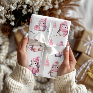 May include: White gift wrap featuring a repeating pattern of penguins wearing pink hats and scarves, with pink Christmas trees. The gift is tied with a white satin ribbon and bow. The word "Nicole" is printed on the wrap.