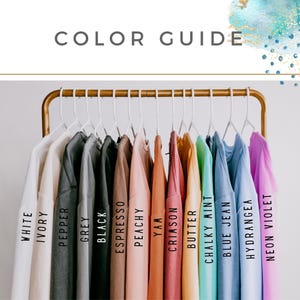 May include: A color guide featuring a variety of t-shirts in different colors, including white, ivory, pepper, grey, black, espresso, peachy, yam, crimson, butter, chalky mint, blue jean, hydrangea, and neon violet. The shirts are hanging on a gold rack.