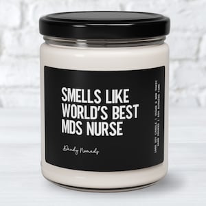 May include: A white candle with a black label that reads "Smells Like World's Best MDS Nurse" and "Dendy Nomads". The label also includes the text "100% Soy Candle | Vegan & Non Toxic | Hand Poured | 60hr Burning Time".