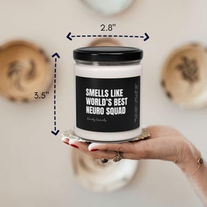 K&ouml;nnte beinhalten: Eine wei&szlig;e Kerze mit einem schwarzen Etikett, auf dem "Smells like world's best neuro squad" steht. Die Kerze ist 3,5 Zoll hoch und 2,8 Zoll breit.