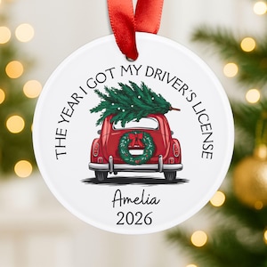 Pu&ograve; includere: Un ornamento rotondo in ceramica bianca con un nastro rosso. Il design presenta un'auto rossa che trasporta un pino verde sul tetto, una ghirlanda sul bagagliaio e il testo THE YEAR I GOT MY DRIVER'S LICENSE, Amelia e 2026 stampato in nero.