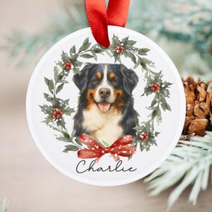 May include: A white, round Christmas ornament featuring a Bernese Mountain Dog portrait surrounded by a holly wreath and a red bow. The dog's name, "Charlie," is written in script below. A red ribbon is attached for hanging.