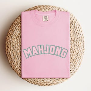 May include: A light pink t-shirt with the word "MAHJONG" in a teal and white arched font. The shirt is folded and rests on a round, woven, light brown surface.