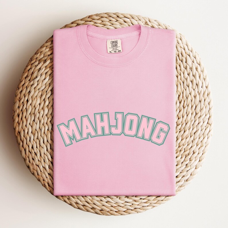 May include: A light pink t-shirt with the word "MAHJONG" in a teal and white arched font. The shirt is folded and rests on a round, woven, light brown surface.