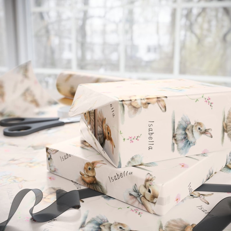 May include: A stack of gift wrap featuring a pink background with a repeating pattern of watercolor bunnies wearing tutus. The text "Isabella" is printed on the top sheet of wrapping paper.
