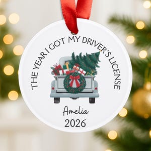 Pu&ograve; includere: Un ornamento rotondo in ceramica bianca con un nastro rosso. Il design raffigura un pickup azzurro che trasporta un albero di Natale e regali, con una ghirlanda sul portellone. Il testo recita: THE YEAR I GOT MY DRIVER'S LICENSE, Amelia, 2026.