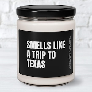 May include: A white candle with a black label that reads "Smells Like A Trip To Texas".