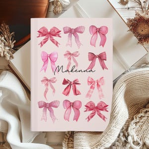 May include: Pink notebook with the name Makenna written in black cursive. The notebook is decorated with pink watercolour bows.