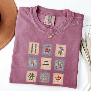 May include: A mauve Comfort Colors t-shirt featuring a nine-tile Mahjong design. The tiles are in shades of green, blue, and red. A gold necklace with a pendant rests on the shirt. The shirt is size S-P-CH.
