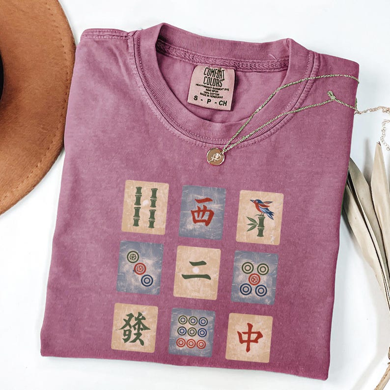 May include: A mauve Comfort Colors t-shirt featuring a nine-tile Mahjong design. The tiles are in shades of green, blue, and red. A gold necklace with a pendant rests on the shirt. The shirt is size S-P-CH.