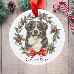 May include: A white, round Christmas ornament featuring a portrait of a dog within a wreath of green leaves and red berries. A red bow and the name "Charlie" are at the bottom. A red ribbon is attached for hanging.