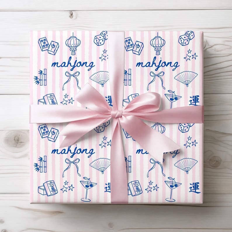 May include: Gift-wrapped present with pink ribbon. The wrapping paper features a pattern of blue Mahjong tiles, dice, lanterns, fans, and text on a pink and white striped background. The word "mahjong" is written in blue.