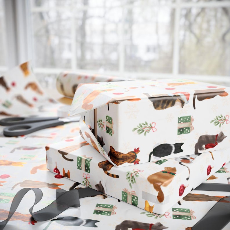 K&ouml;nnte beinhalten: Zwei Geschenkboxen, verpackt in wei&szlig;em Papier mit einem sich wiederholenden Muster von Katzen in verschiedenen Farben und Posen. Die Boxen sind mit gr&uuml;nen Geschenkboxen, roten Kugeln und Zweigen verziert. Eine Rolle passendes Geschenkpapier und eine Schere sind sichtbar.